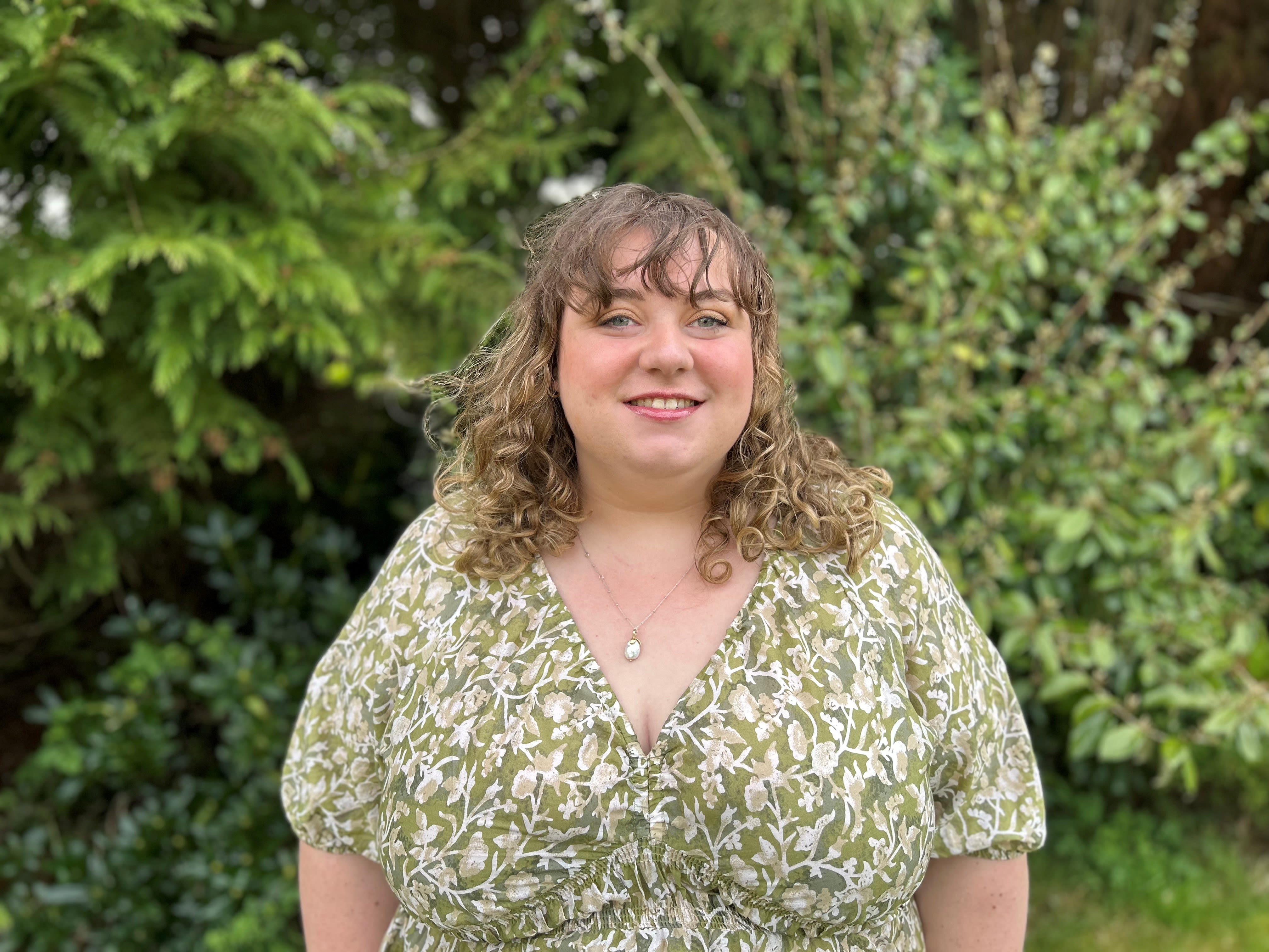 Georgia Horsell smiles warmly wearing a green patterned dress, while standing in front of greenery 