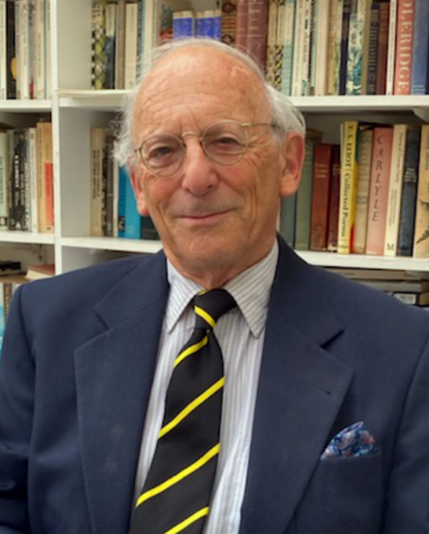 Michael smiles in front of a bookshelf. He is wearing a black and yellow striped tie, a white shirt and a navy blue suit jacket.