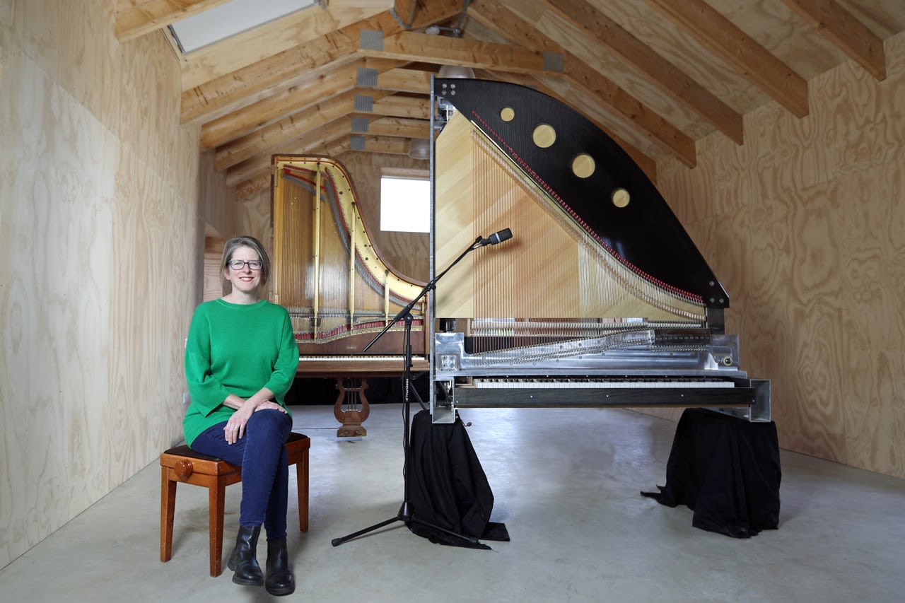 Sarah sits smiling in a green jumper in front of two of her vertical pianos.