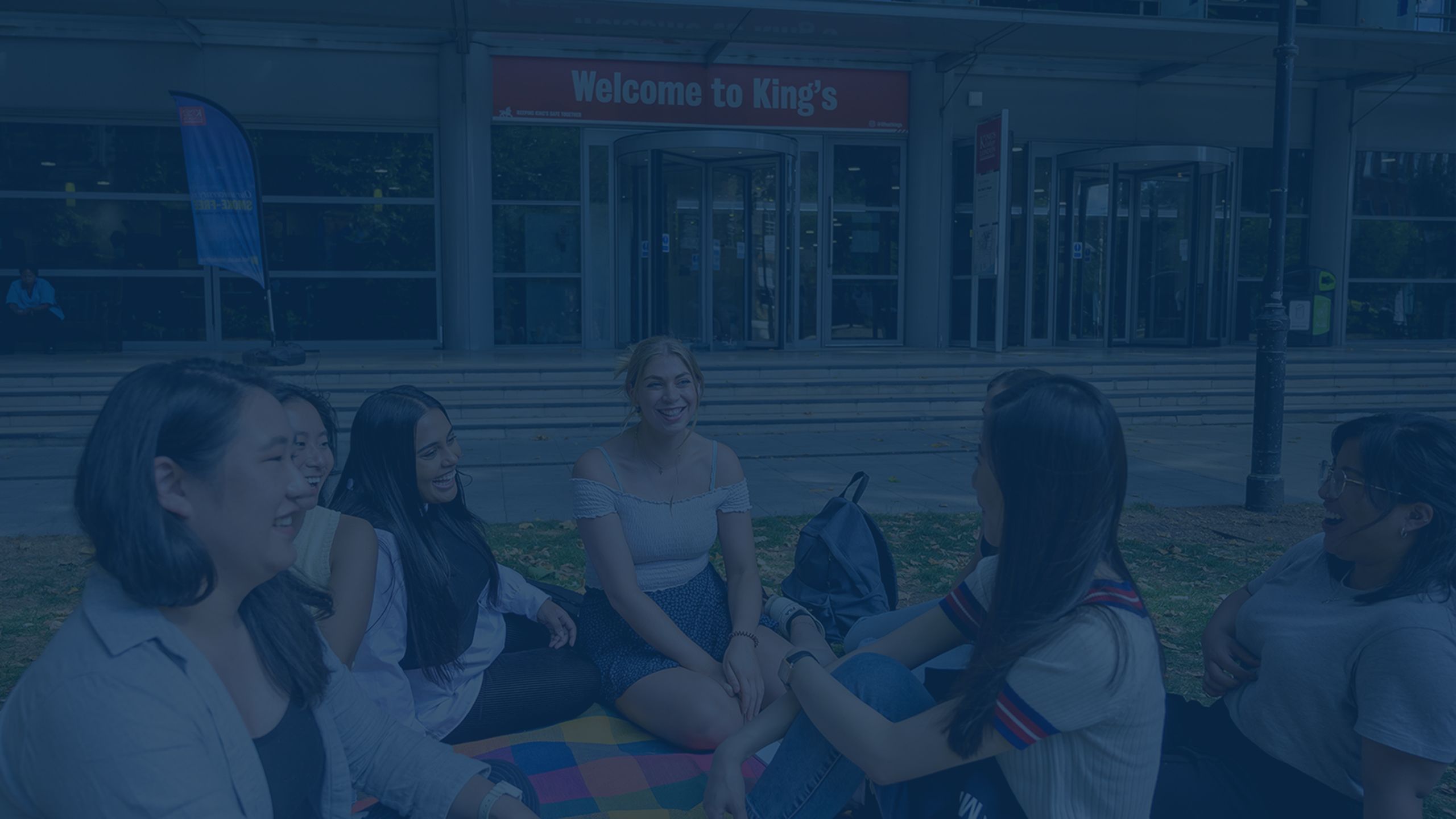 The same image as before of students sitting on a blanket on the quad at Guy's campus. This time the image is overlaid in dark blue.