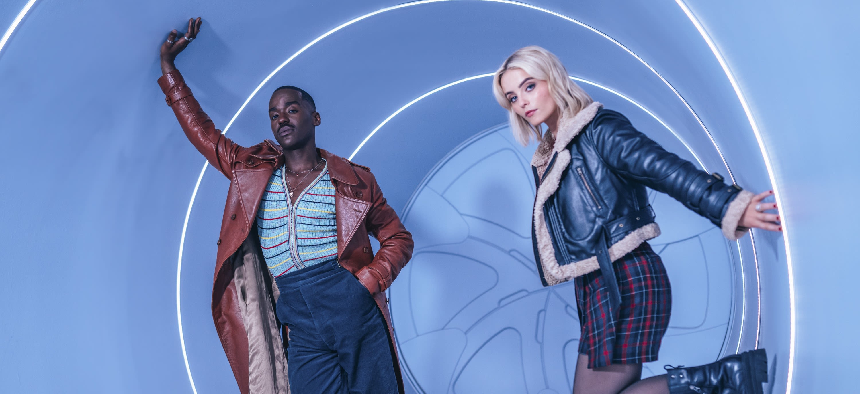 A still image of Ncuti Gatwa and Millie Gibson as their characters in Dr Who. They stand inside a blue tunnel, holding on to the sides. 