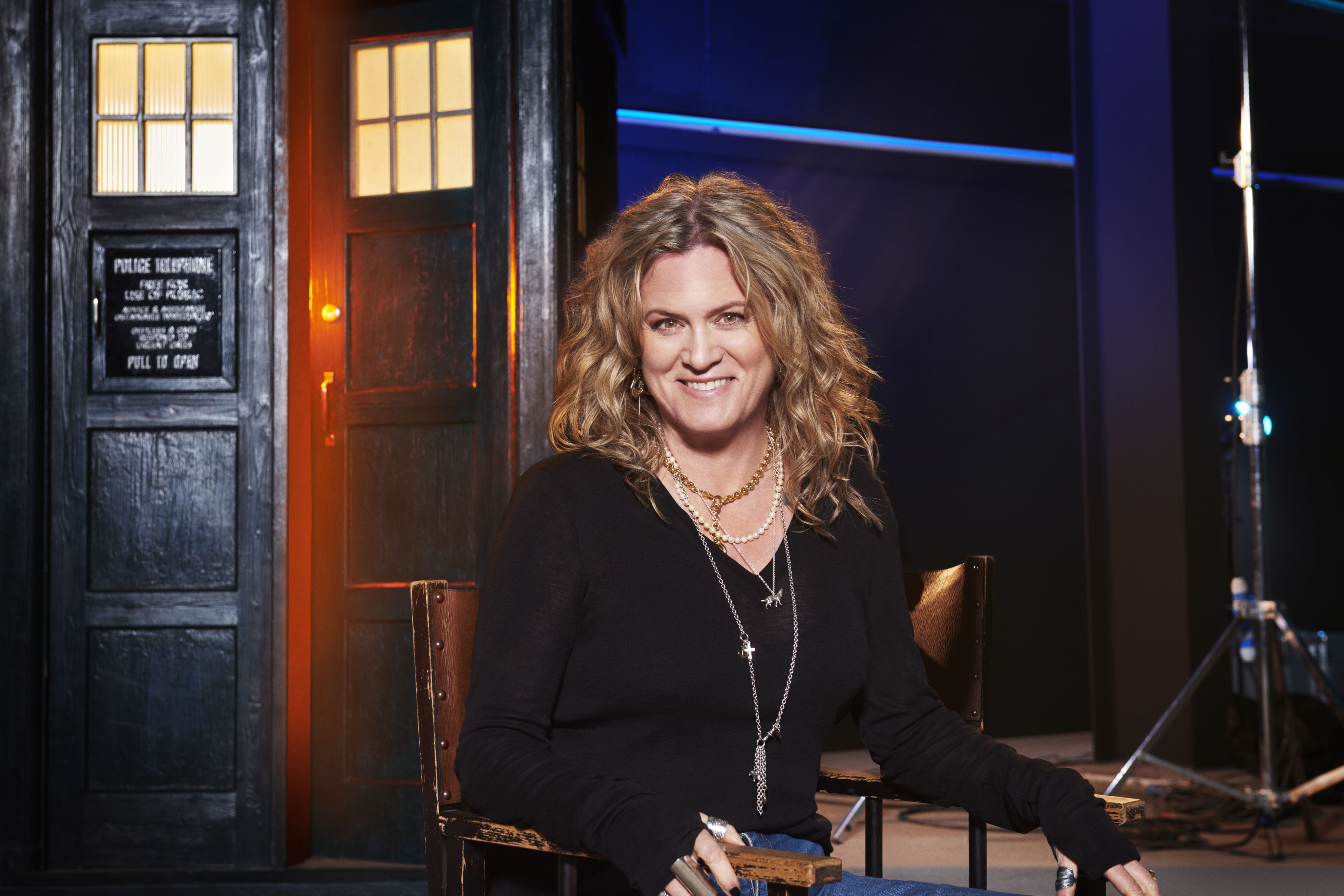 Jane is pictured sitting in an old chair in front of the Doctor Who Tardis. She has shoulder length blonde hair and she is wearing a black top, blue jeans and a long silver necklace.