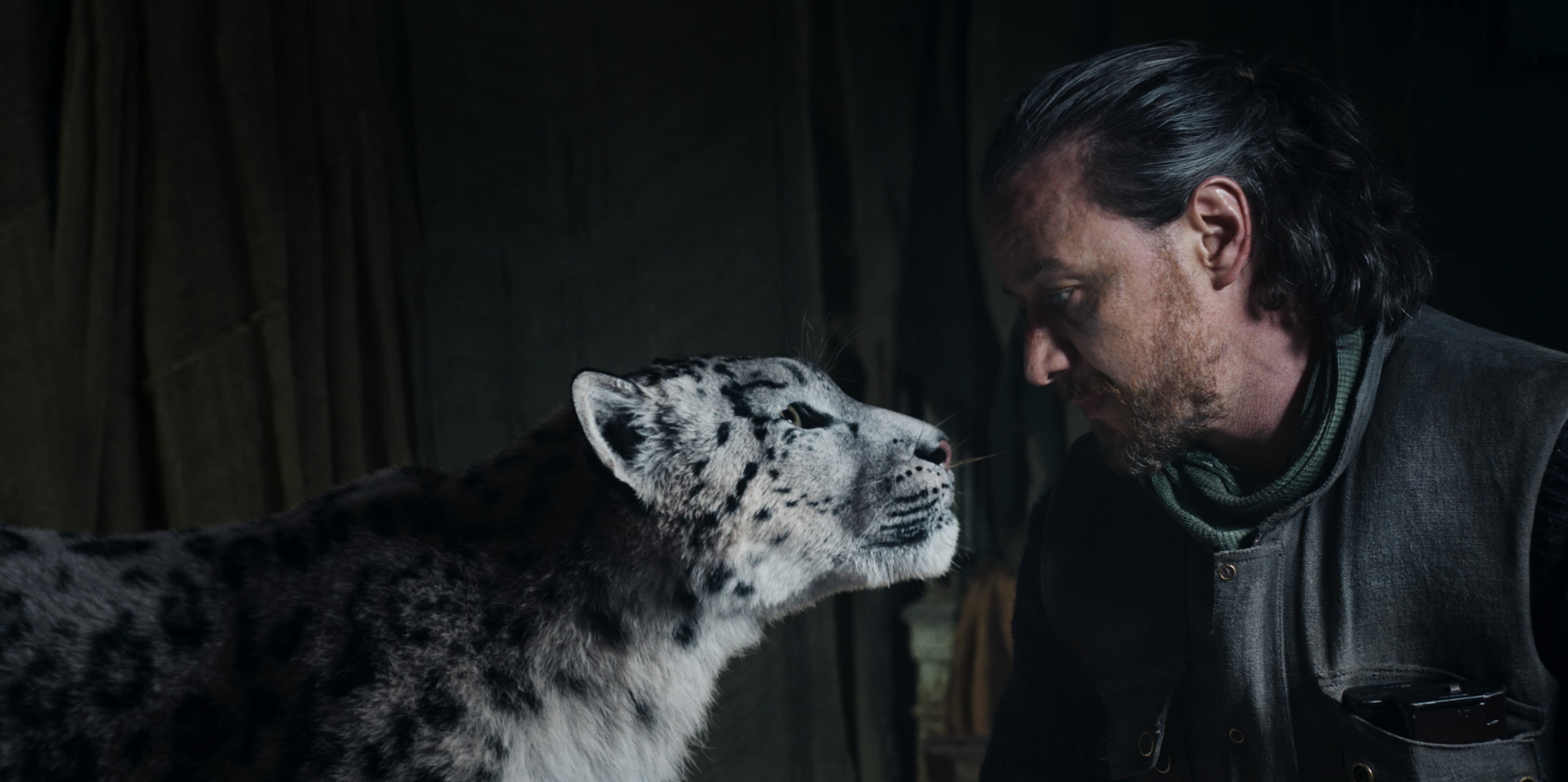 A man with long tied back black hair stares into the eyes of a snow leopard. The image is a production still from 'His Dark Materials' 