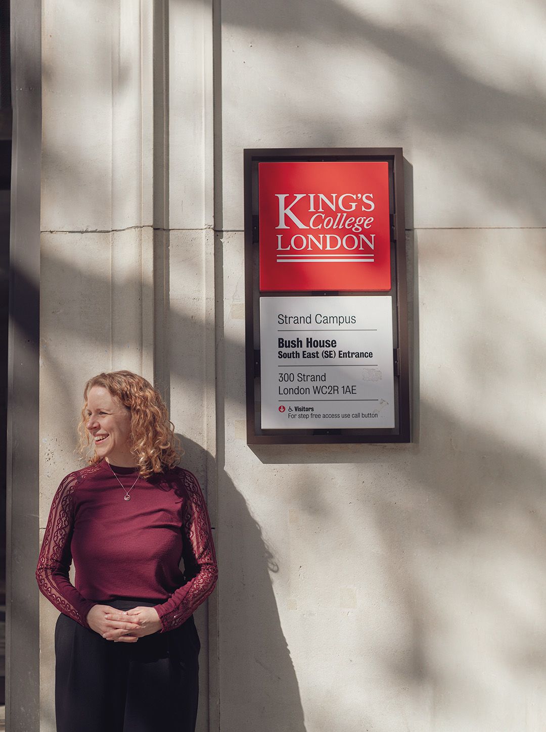 Emma Keith looking into the distance smiling while standing next to a campus sign on Bush House.