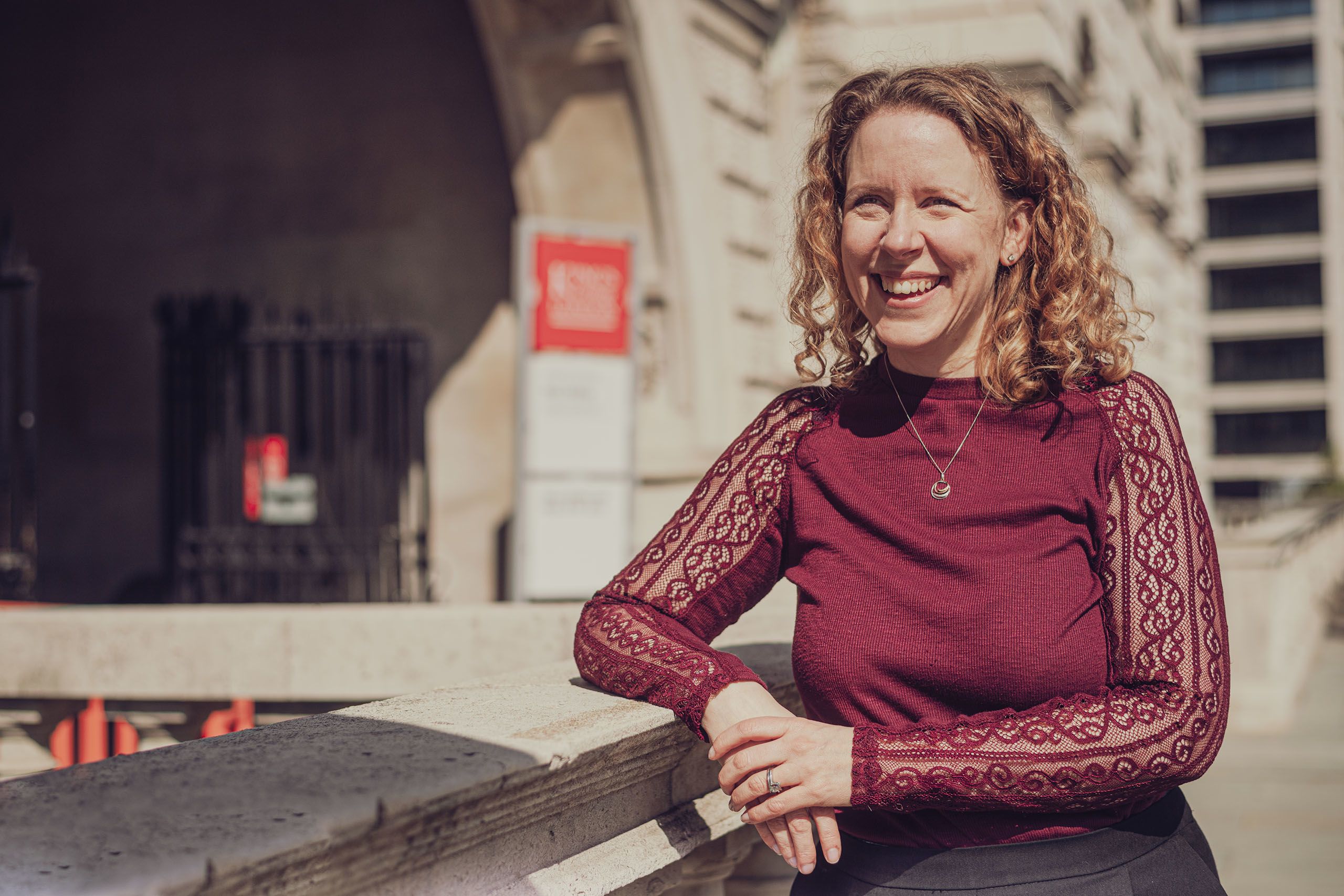 Emma stands smiling on a sunny day, leaning against a stone wall. 