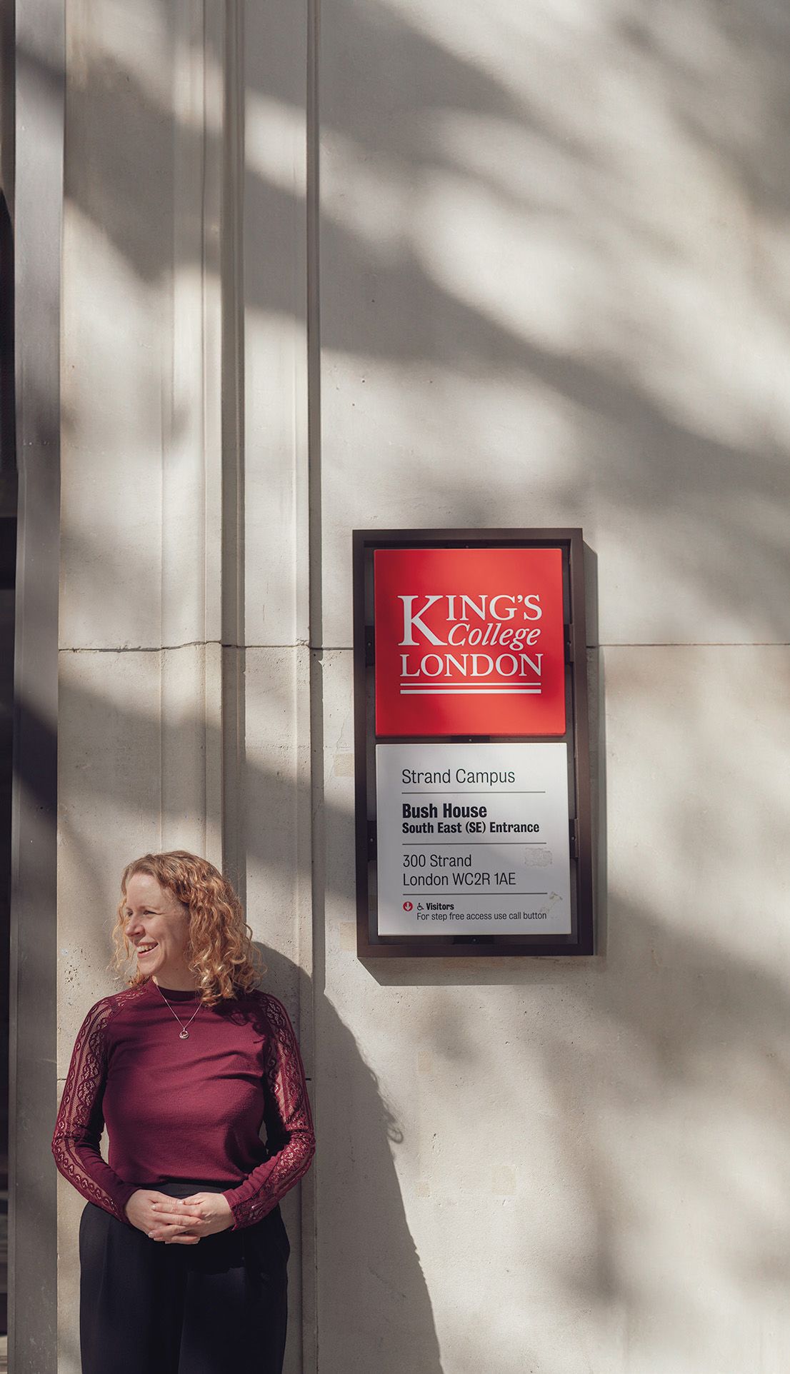 Emma Keith looking into the distance smiling while standing next to a campus sign on Bush House.