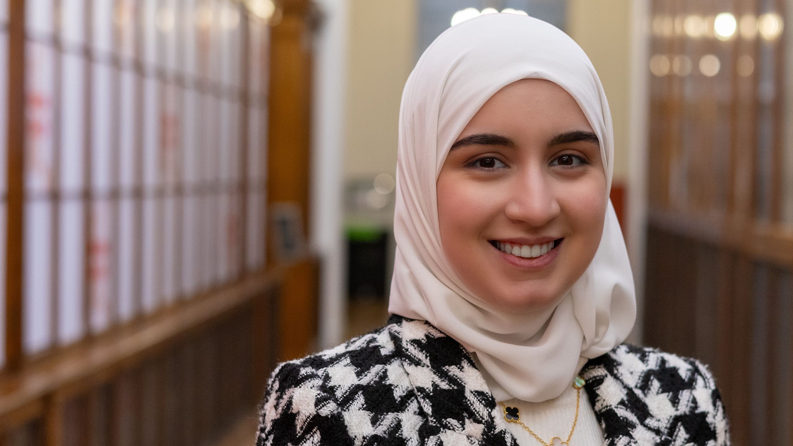 Baian smiles warmly. She is wearing a black and white dog tooth coat and a white head scarf.