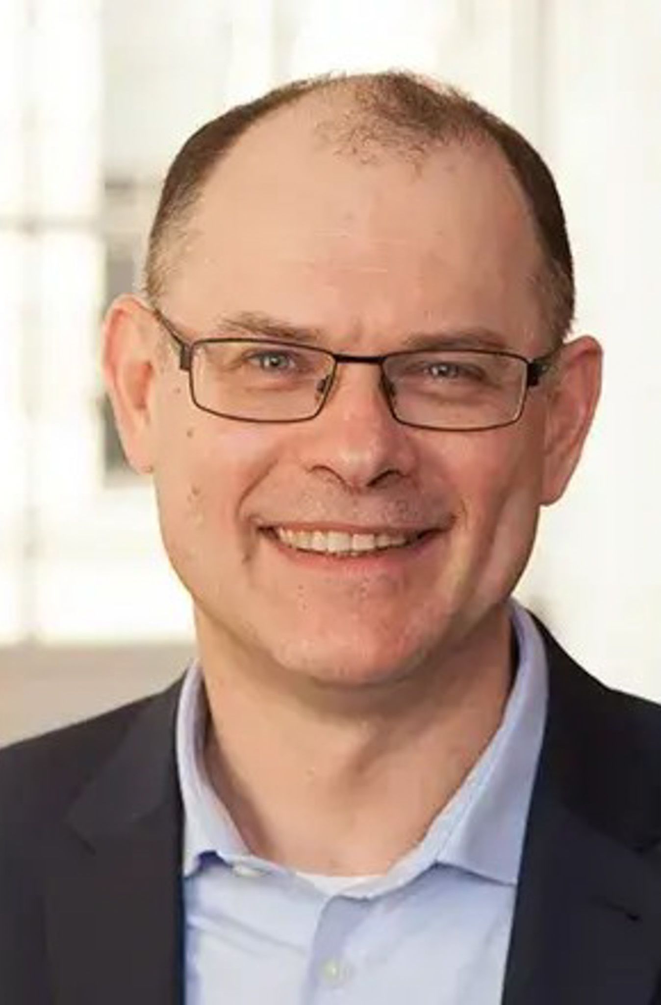 Alexander Türk, a middle-aged man with short hair and glasses, smiling while wearing a dark blazer over a light shirt. The background is softly blurred.