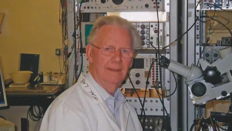 Thomas Sears with grey hair wearing a lab coat, standing in a lab filled with equipment, looking at the camera.
