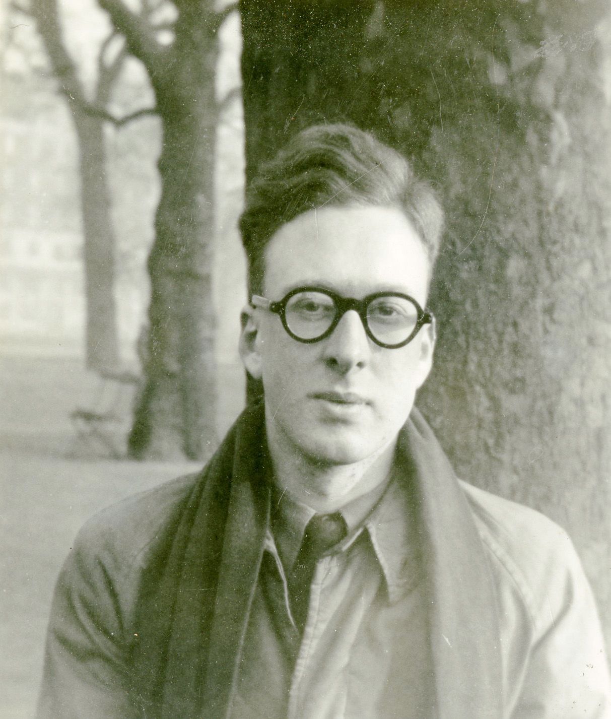 A picture of Professor Peter Higgs. He is pictured looking solemly down the lens. The picture is from the 1950s. He is wearing a coat, a scarf, a thick rimmed glasses. The picture is in sepia