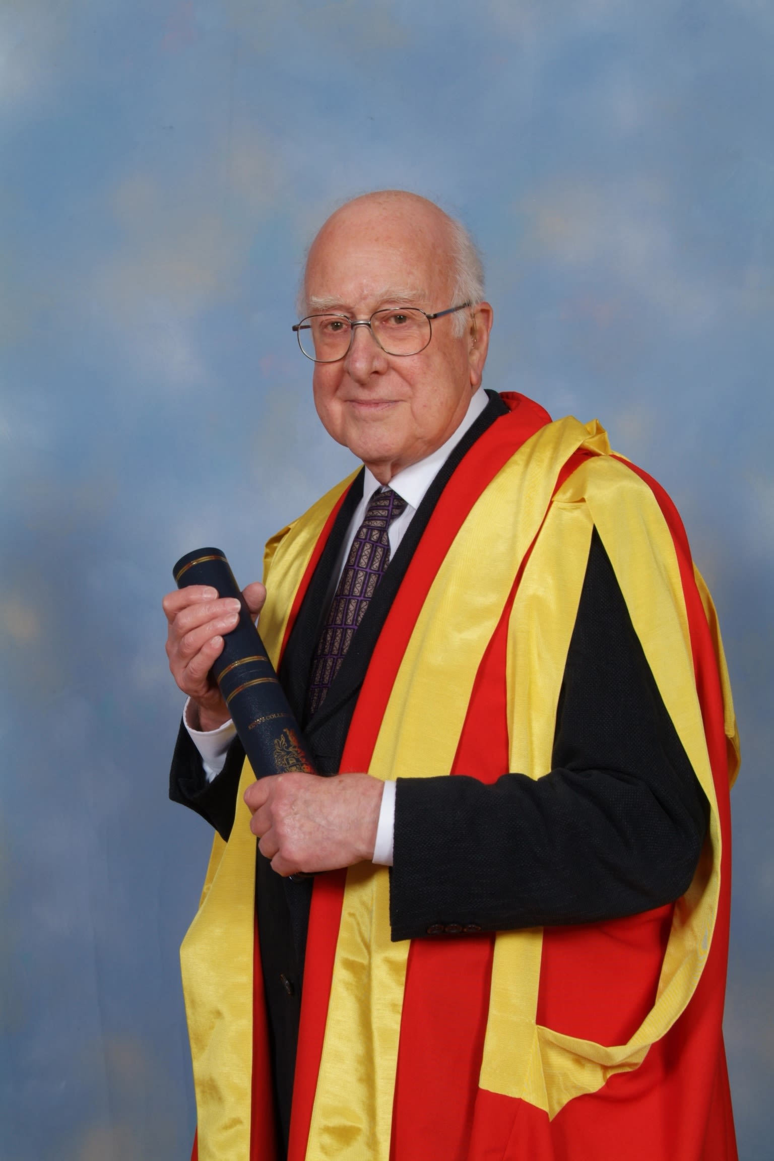 Professor Higgs is pictured in a dark suit and yellow and red academic dress. He clutches a scroll. 