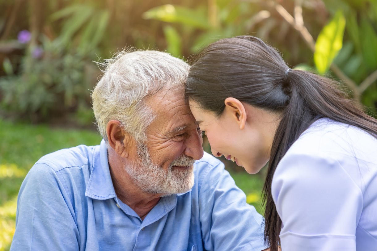 Smiling nurse and eldery senior man touching heads in garden in nursing home