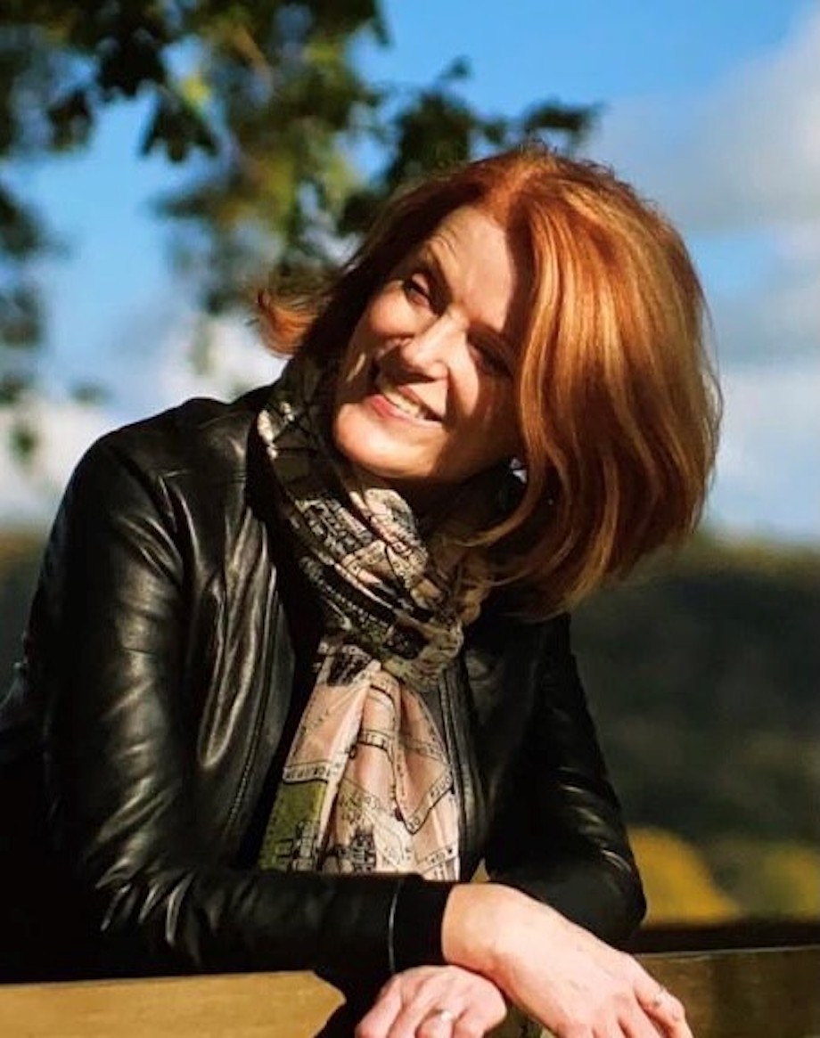 A woman outside on a sunny day, leaning on a gate, smiling and wearing a scarf and leather jacket