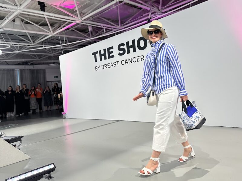 A woman on the catwalk at the Breast Cancer Now Show, smiling and wearing a summer hat, sunglasses and a striped shirt