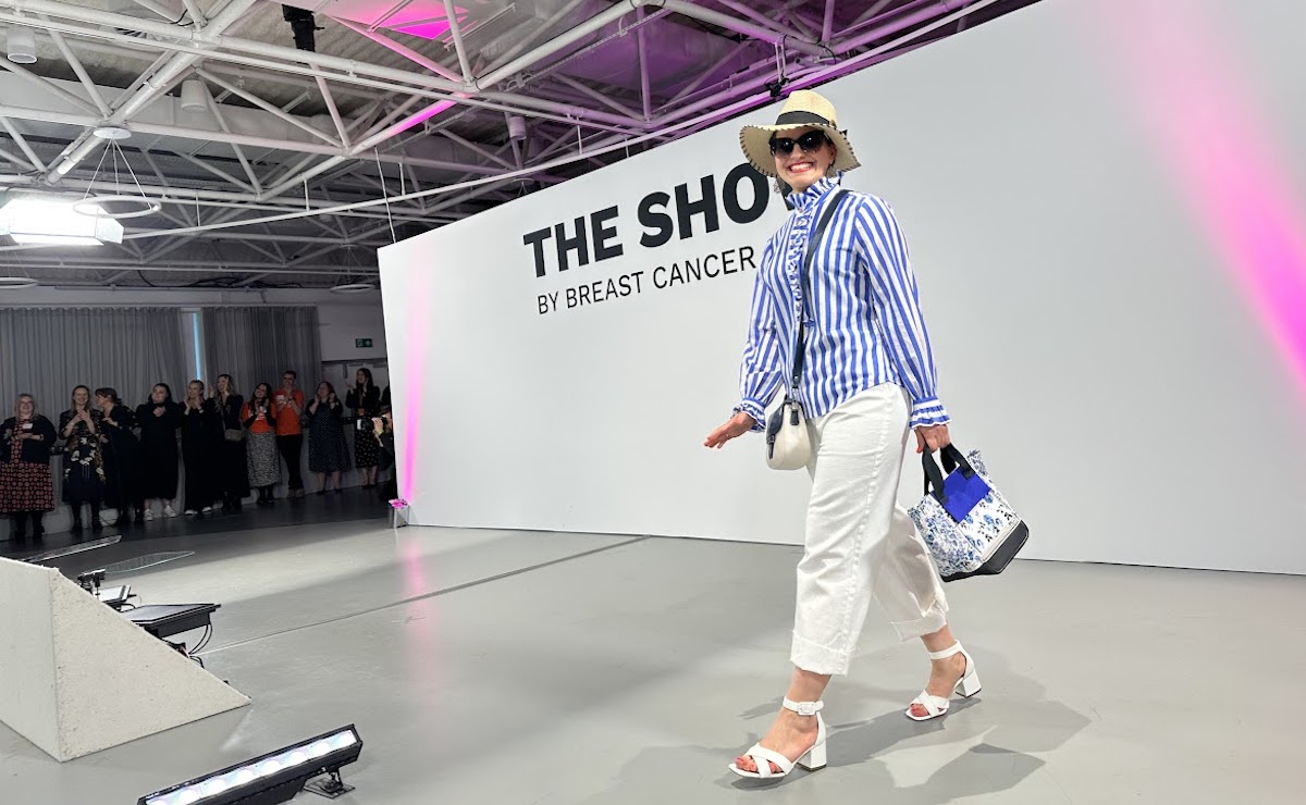 A woman on the catwalk at the Breast Cancer Now Show, smiling and wearing a summer hat, sunglasses and a striped shirt