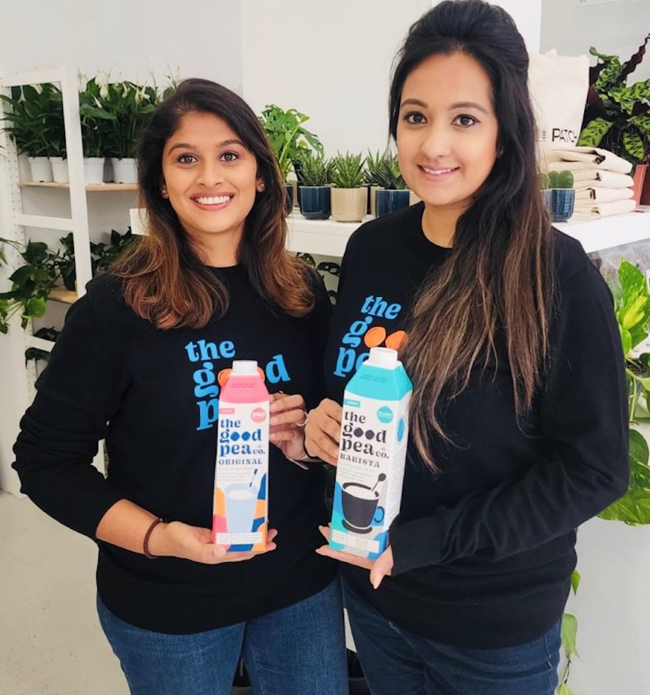 Two young women holding 'The Good Pea Co' milk cartons and smiling, with potted plants in the background