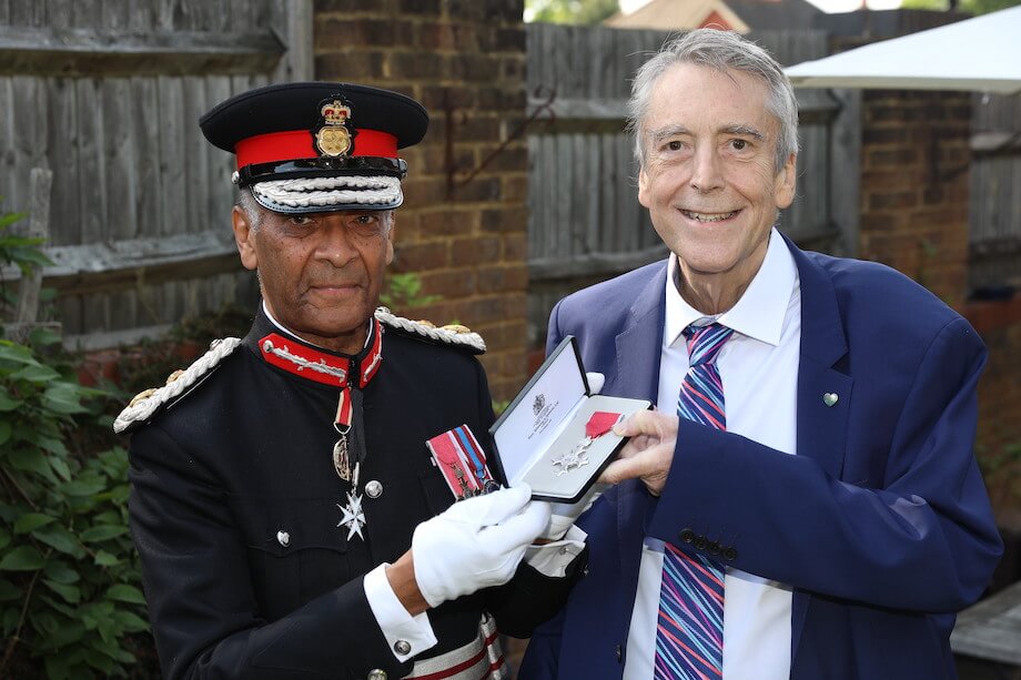 A man in a suit and tie receiving an MBE medal from a man in a military hat and uniform, smiling in a garden