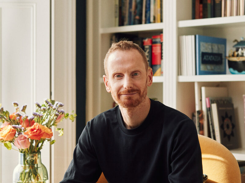 Headshot of a young man with a beard in a black jumper, smiling in front of bookshelves, next to a vase of flowers