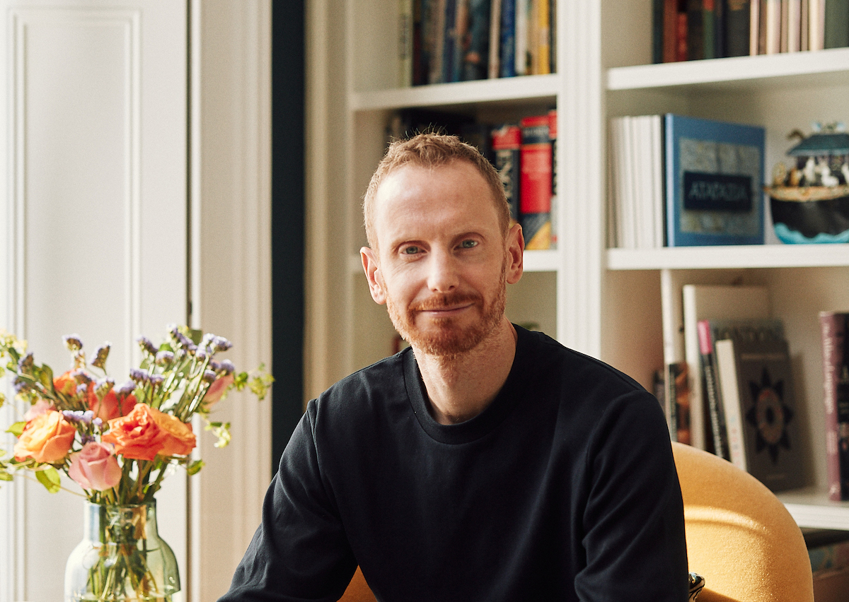Headshot of a young man with a beard in a black jumper, smiling in front of bookshelves, next to a vase of flowers