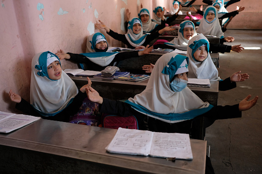 Young Afghan girls in a classroom with their arms outstretched, smiling, wearing blue and white Hijabs