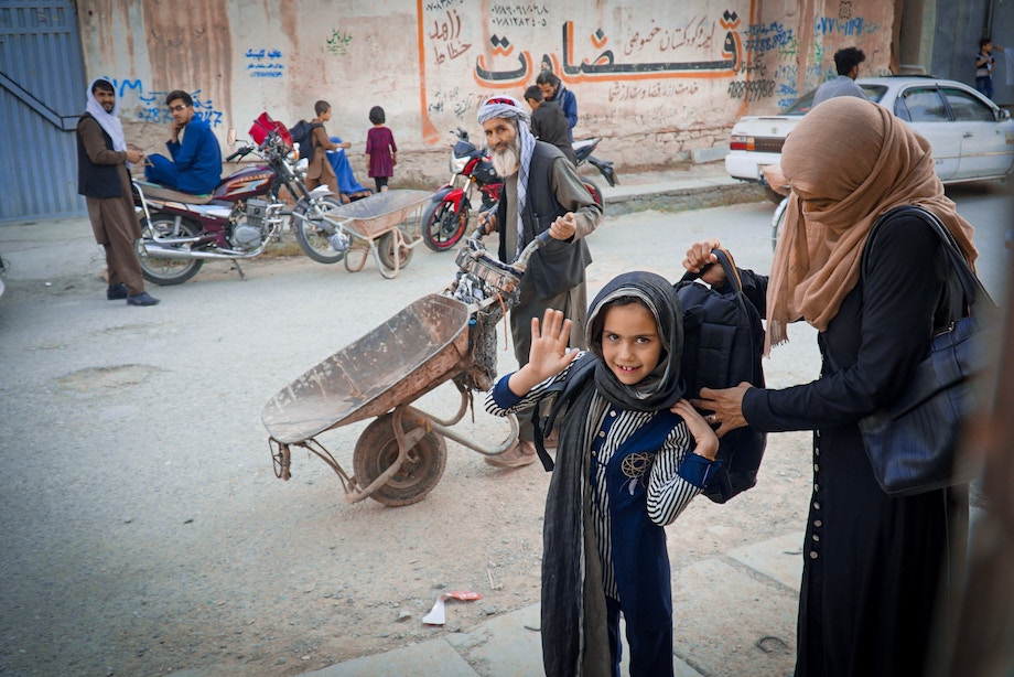 Street photo in Afghanistan of a mother putting a backpack on child, who is smiling and waving at the camera. A man pushes a wheelbarrow in the background