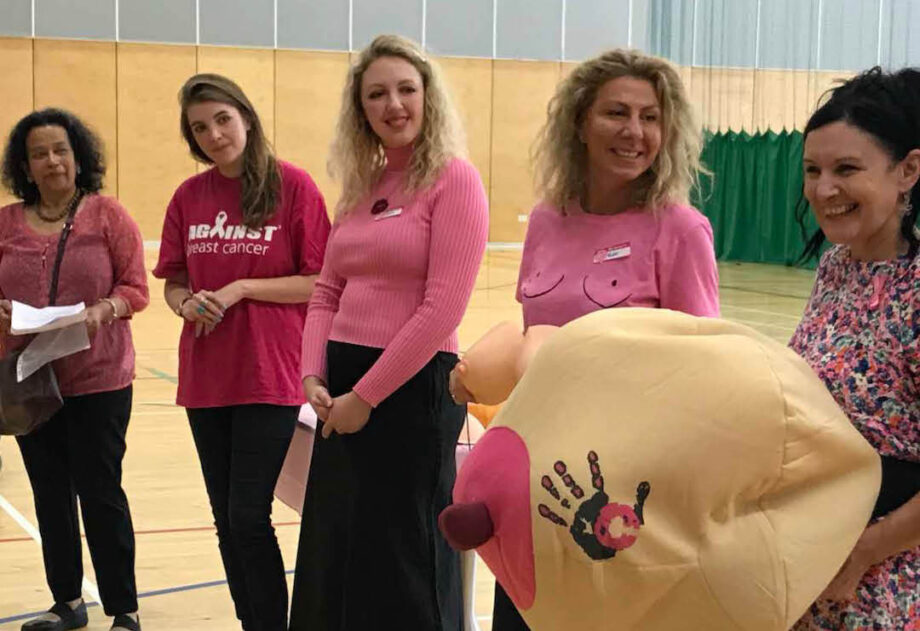 A group of women in a gym hall, wearing pink and laughing. One is holding an inflatable breast.