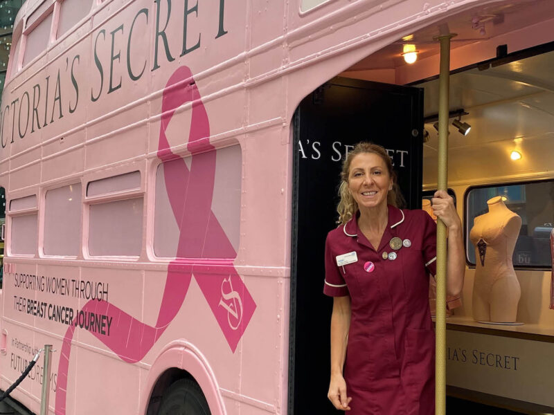 Outside shot of a woman in a nurse uniform standing on the bottom deck of a pink Breast Cancer bus
